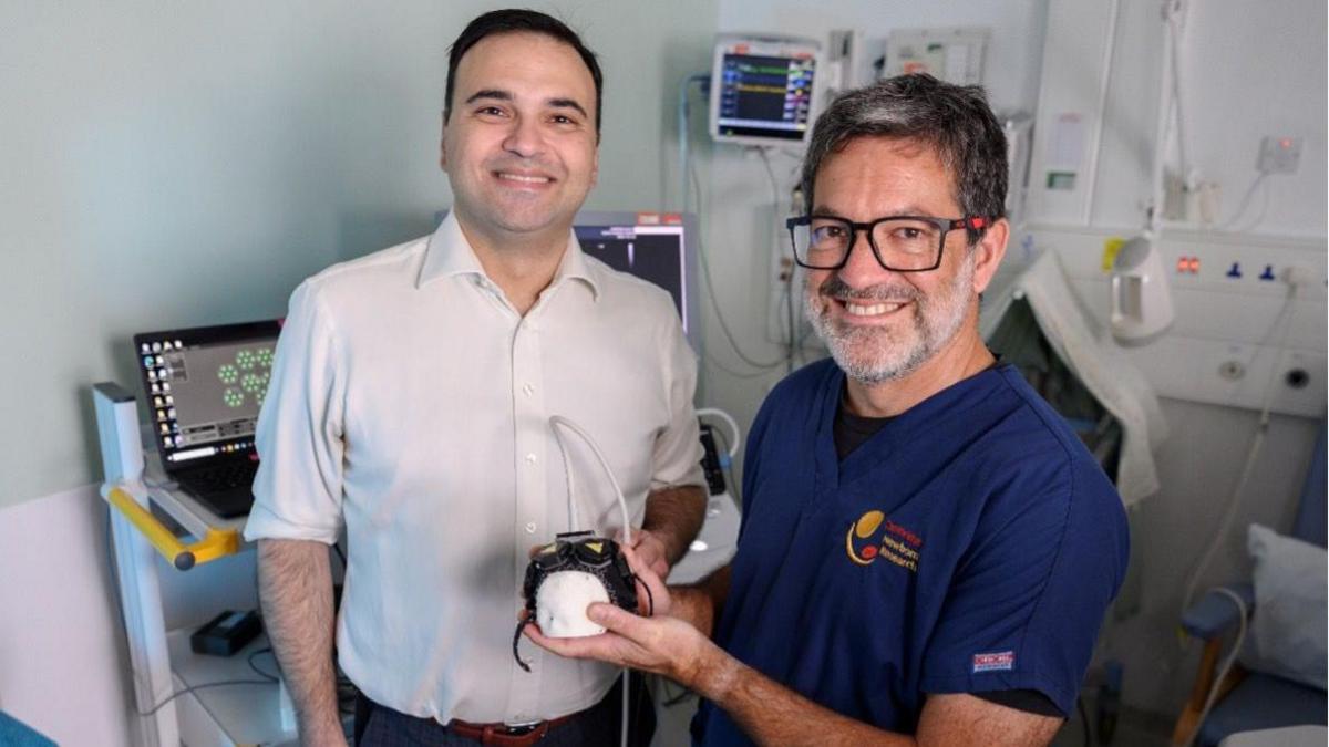 Alexis Joannides is on the left of the picture wearing a white shirt and smiling at the camera. Topun Austin, is standing to his right wearing blue hospital scrubs, glasses and is also smiling. He is holding the black sensor cap which is fitted onto a small polystyrene model of a head. They are both in a clinical room with monitors and medical equipment in the background.
