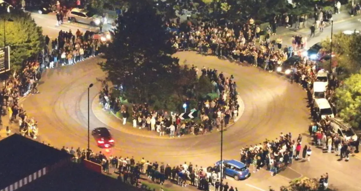 An overhead shot at night time of cars going around a roundabout whilst crowds line the pavement.