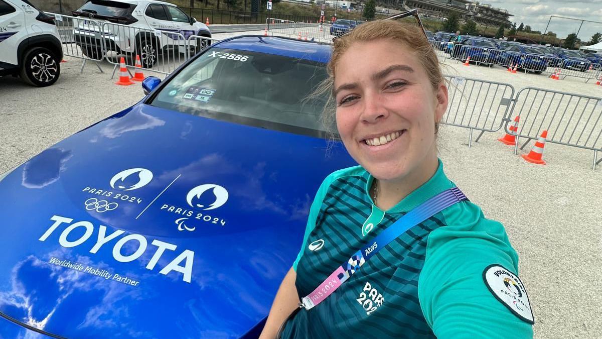 A young woman takes a selfie as she stands in front of a dark blue saloon car with Paris 2024 logos and Toyota branding on its bonnet. She's wearing an official volunteer uniform. In the background about two dozen similar cars are parked in a makeshift carpark.