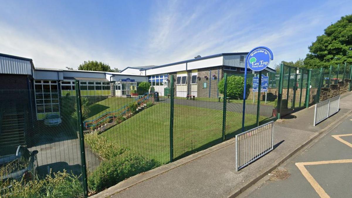 A school with a sloped grass area in front and it is all surrounded by a wire fence. There is a circular blue sign in front. 