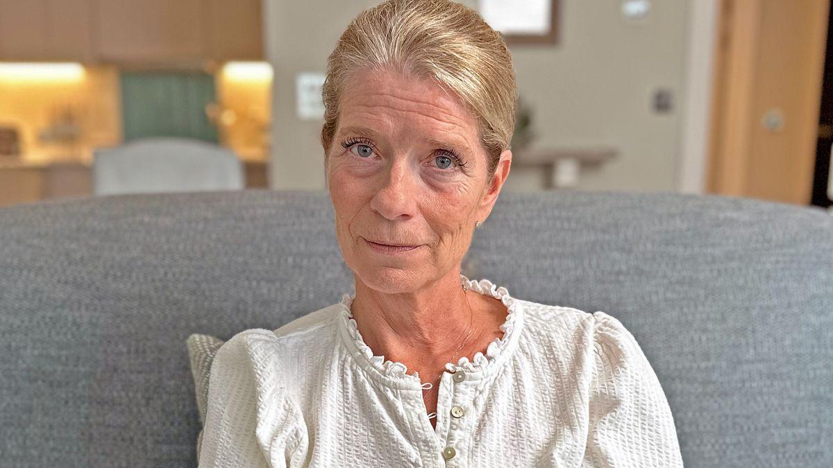 Frances, a middle aged woman, with blond hair, looking directly at the camera. She is wearing a white poplin blouse and is sitting on a plain grey sofa.