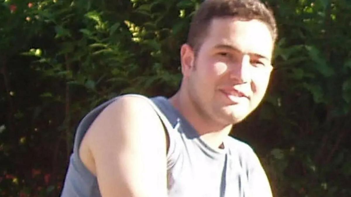 Head and shoulders shot of Jean Charles, looking at the camera, smiling. He is wearing a light coloured sleeveless top and is in front of some hedging.