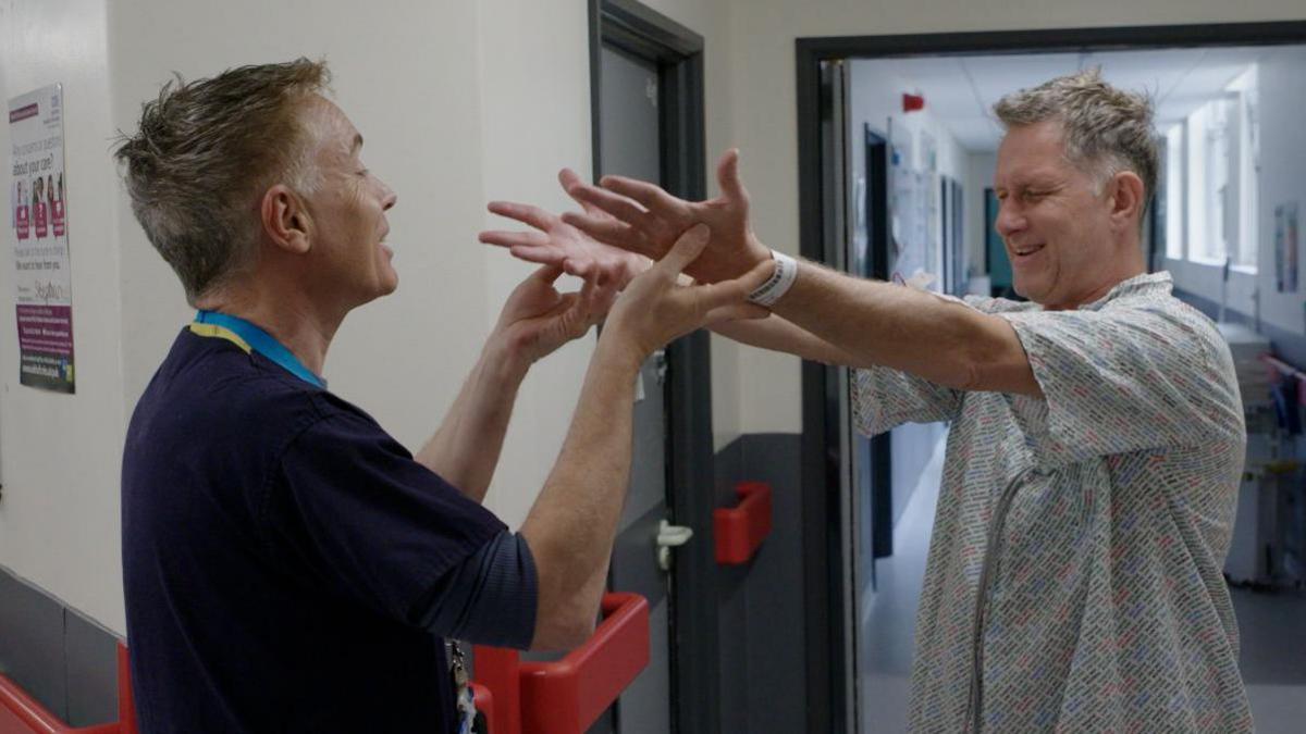 A man in a hospital gown smiles as he lifts his hands with the aid of a doctor 