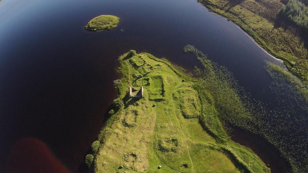 A drone image showing archaeological remains at Finlaggan. There are ruined walls and rectangular shapes beneath grass-covered ground on an area of land that sticks out into the loch. The loch's waters are a peaty brown.