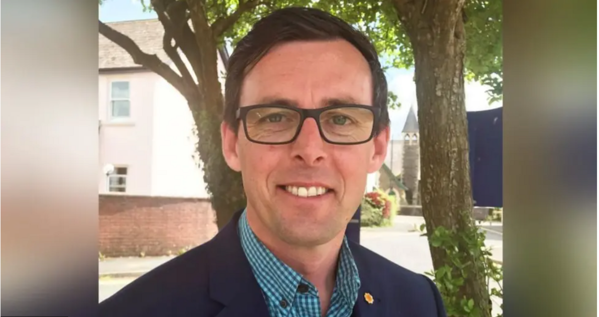 Darren Price yn edrych tuag at y camera gyda choed ac adeilad gwyn yn y cefndir. Mae ganddo lygaid glas, gwallt brown ac yn gwisgo sbectol.