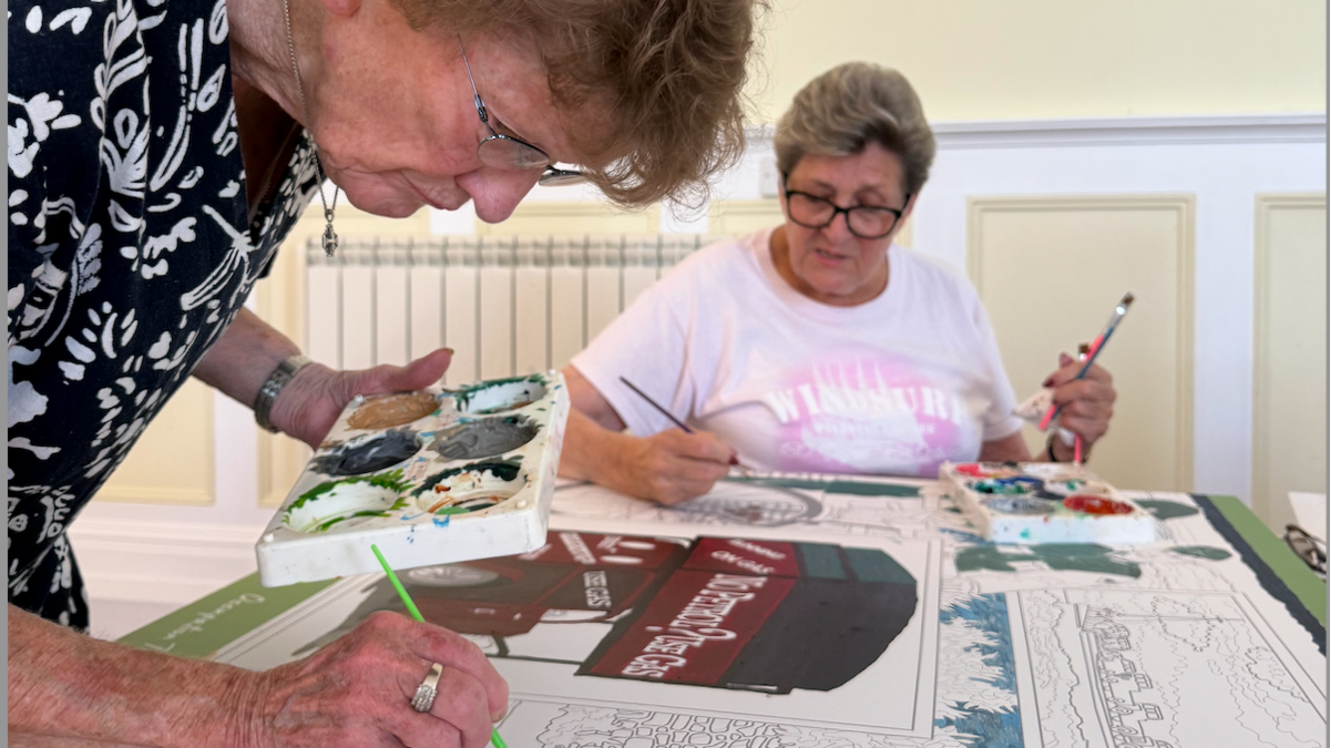 The image shows two people working on a large canvas that has an outlined design ready for painting. The design includes detailed illustrations of buildings, a horse-drawn cart, and a vintage delivery van with the words “No Petrol? Use Gas” clearly visible. One person is applying paint to part of the scene using a brush and a palette, while the other is seated and holding a paintbrush, appearing to work on another section. The canvas is spread out on a table, and several paint colours are visible on palettes.
