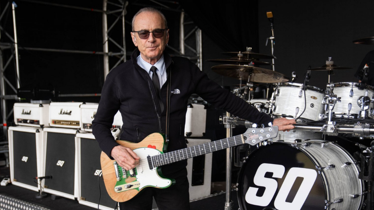 Franics Rossi stands in black hoodie with a white shirt and black tie. He holds an electric guitar with all the band equipment behind him.