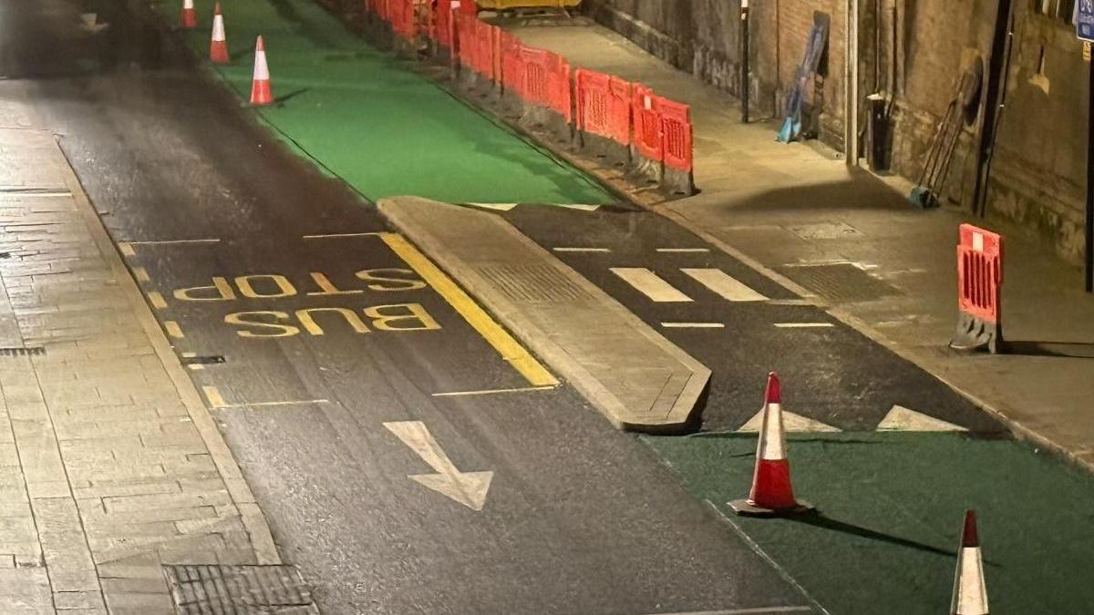A single lane road with a bus stop in the middle. Alongside it is a green cycle lane with a narrow slabbed island in the middle. Traffic cones are being used to separate the road and cycleway.