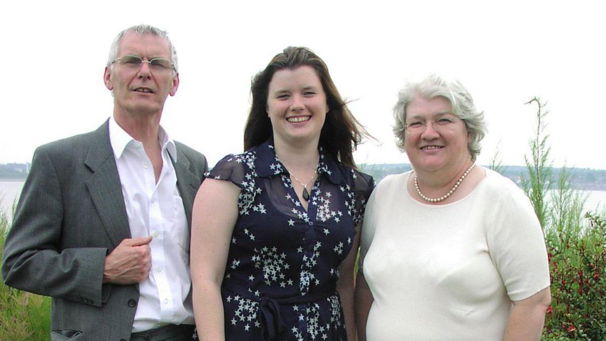 A young Hannah with brown long hair smiles at the camera, standing in the middle of an older man to her left, and an older woman to her right, 