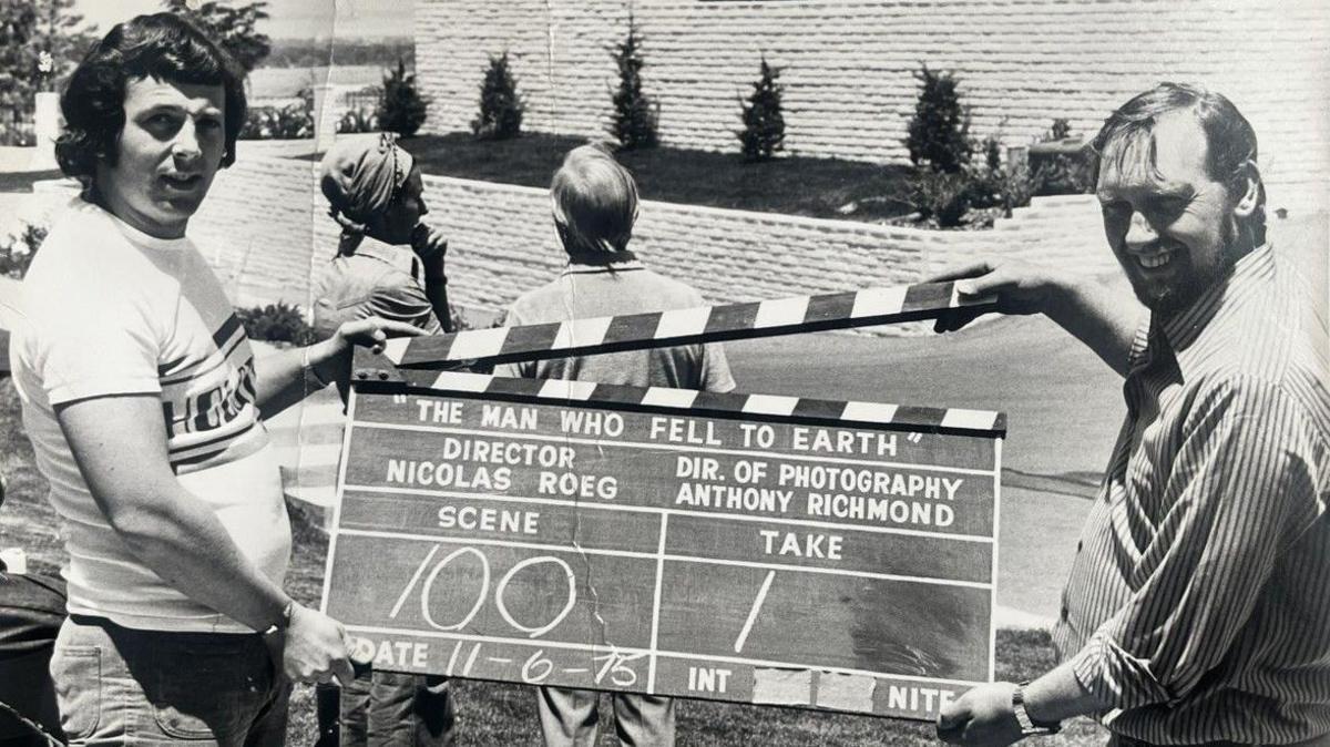 Two men, one on the left and another on the right, are holding a clapperboard, which is in the middle of this black and white photo.