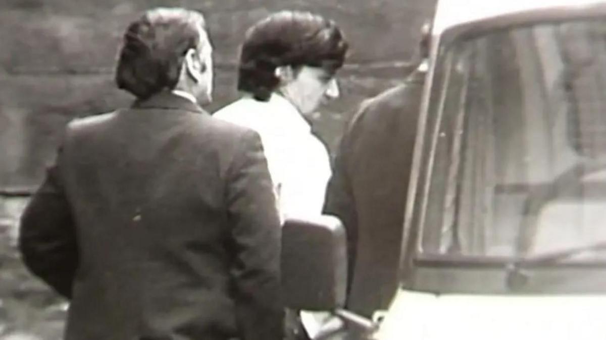 A black and white still from TV footage showing Peter Sullivan, who has thick black hair and is wearing a white shirt, being led to a police van in handcuffs.