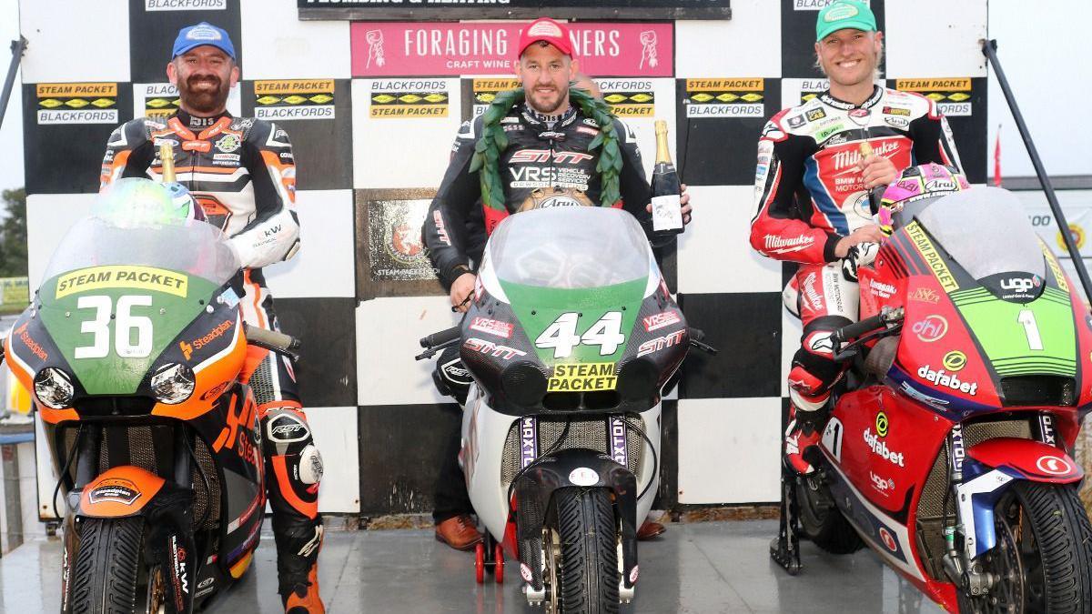 hree riders lined up on their bikes on the podium for the Lightweight race smiling. Rob Hodson is wearing a red cap and black leathers, has a green garland around his neck and is holding a bottle of champagne. His bike is black and white and has the number 44 written in white on a green background on the front. Jamie Coward is on right wearing a blue cap, and orange, black and white leathers, which match the colours of his bike. The number 36 is on the front of the machine in white on a green background. Davy Todd is on the left wearing a turquoise cap and red, white and black leathers, which also match his bike. It has the number one in white with a green background on the front. All three bikes have yellow stickers with the words Steam Packet on them in black lettering.