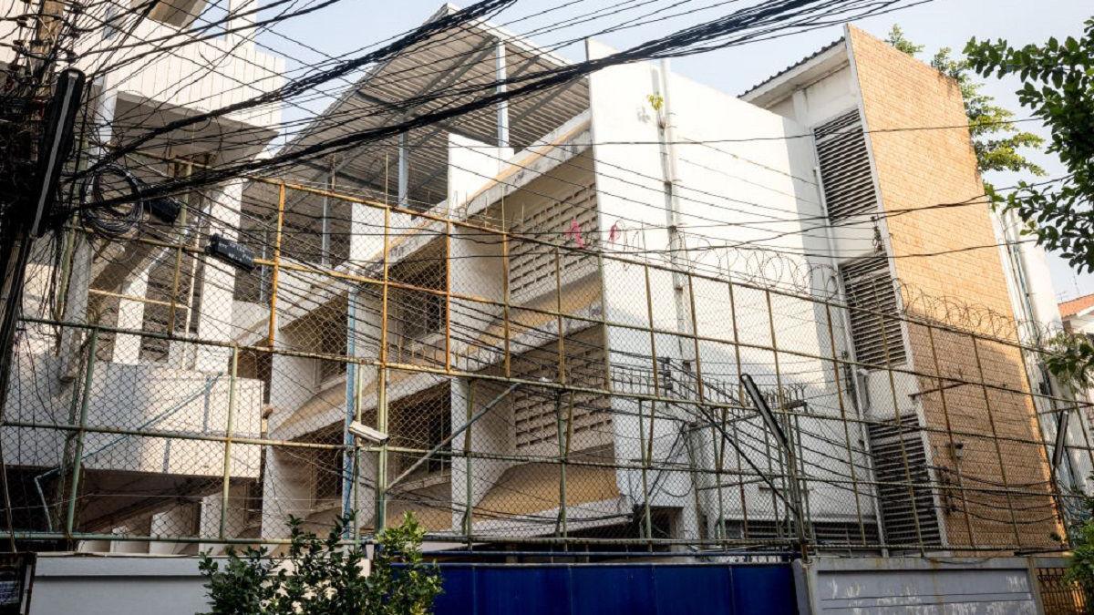 A tall meshed fence topped with barbed wire guards the immigration detention centre where 48 Uyghurs are being held in Bangkok - the building is behind a blue gate and has walls that are white, alongside some built with brown bricks. 