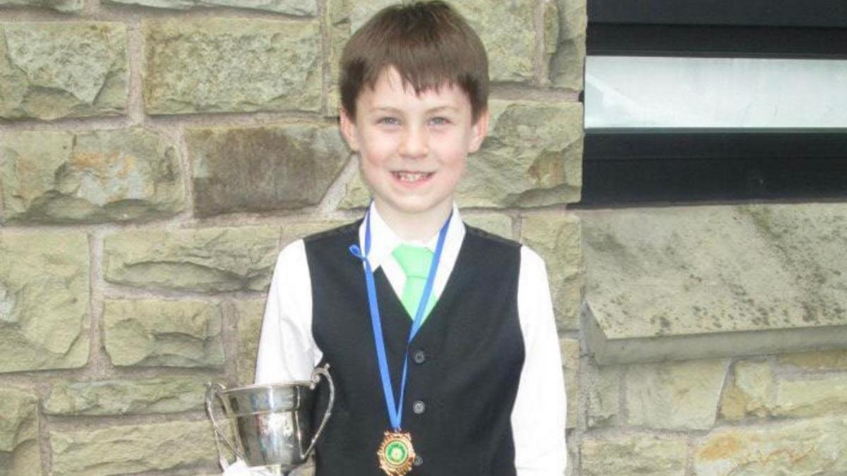 A picture of Barry as a young boy after competing in an Irish dancing competition. He is wearing a white collared shirt, a light green tie and a black waistcoat. He is holding a silver trophy cup and has a gold medal with a blue lanyard on it. He is standing against a light stone wall with some metal vents off to the side.