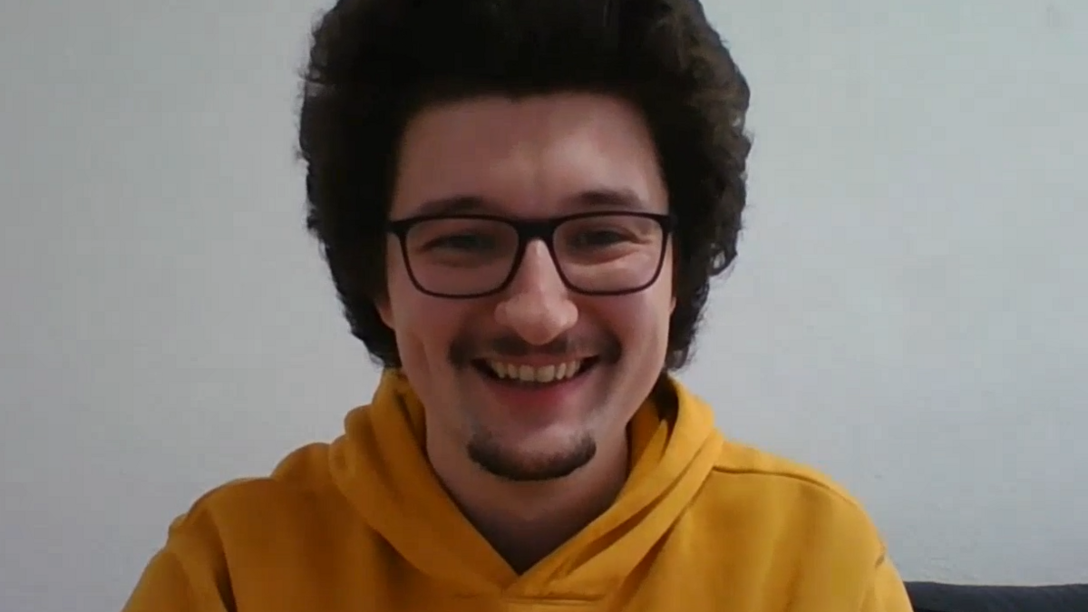 Frank Ilett smiling into a webcam camera. He is wearing a mustard coloured hoodie, black glasses, and has raised long dark hair.