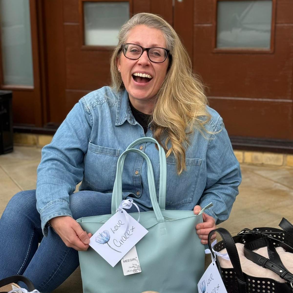 A laughing Hannah O'Callaghan holding a blue handbag with a gift tag on it which reads 'love Grace x'. She has long blonde hair and is wearing a denim shirt.