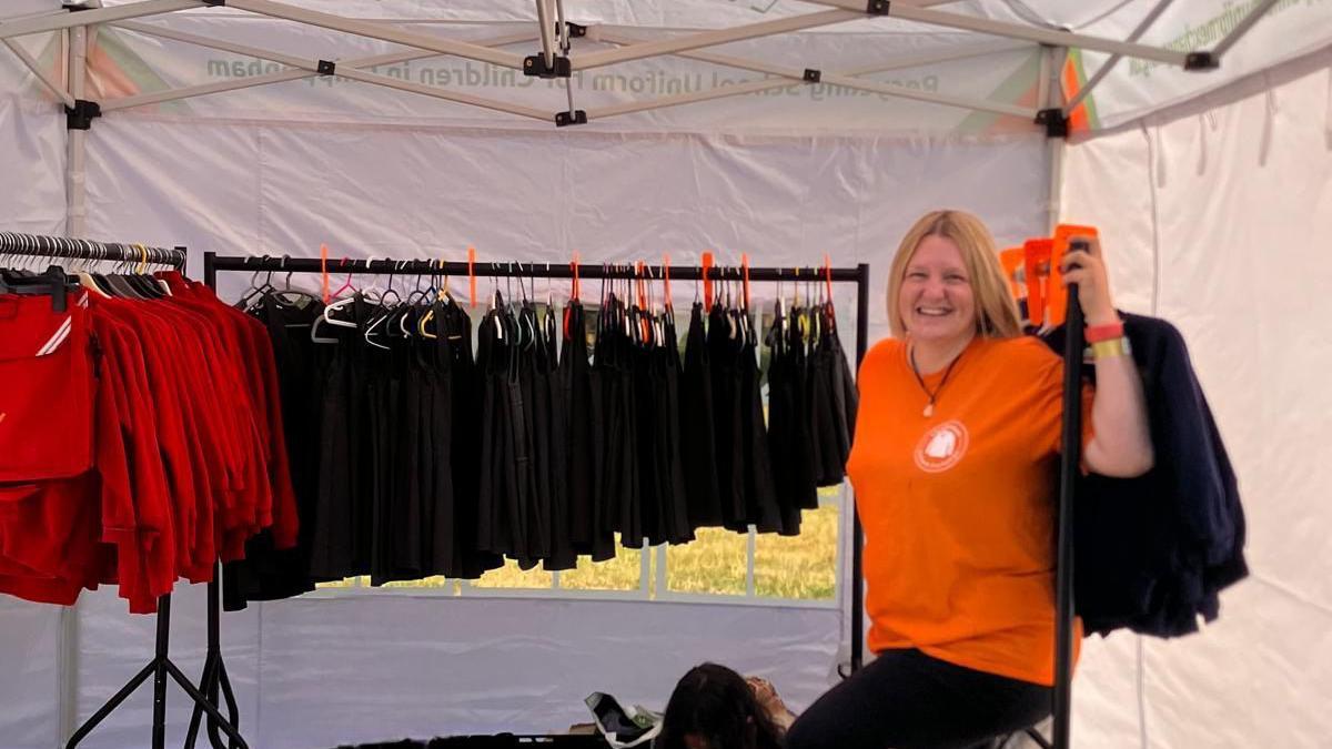 Jade Ugur is smiling and wearing a bright orange T-shirt. She is under a white gazebo with two rails of school uniform.