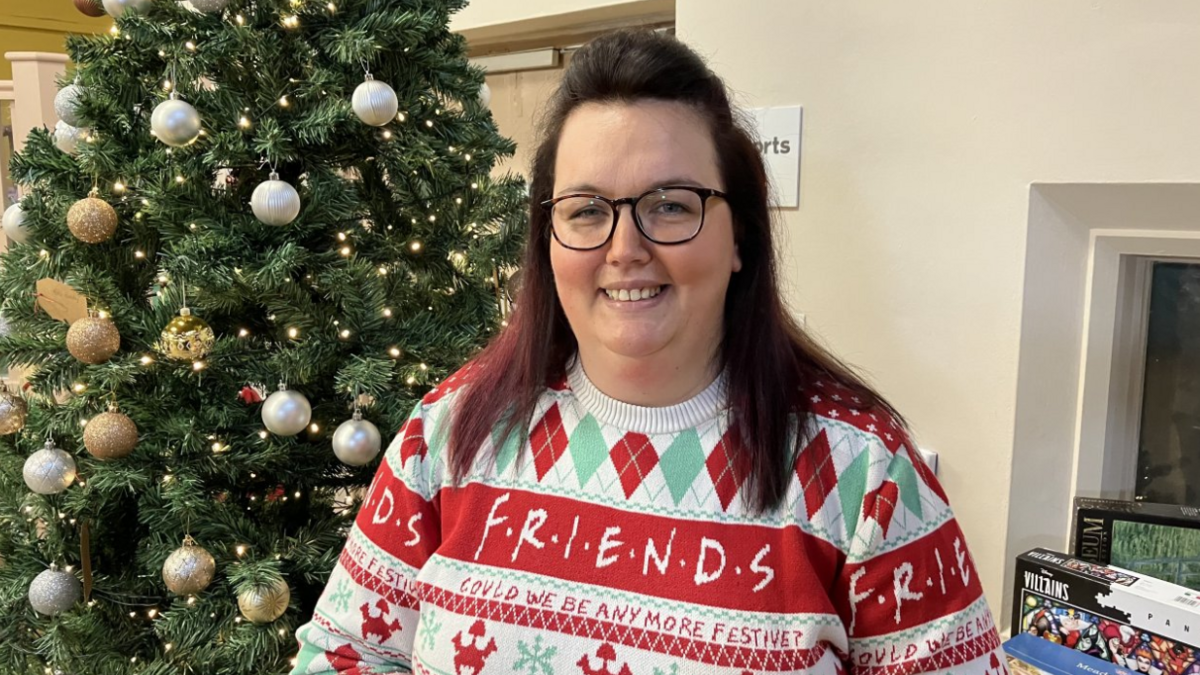 Katie Chun, manager of the Hive, wearing glasses and with brown below-shoulder length hair. She is in a festive Friends Christmas jumper with green and red diamond detail and there is a Christmas tree behind her with silver and gold baubles.