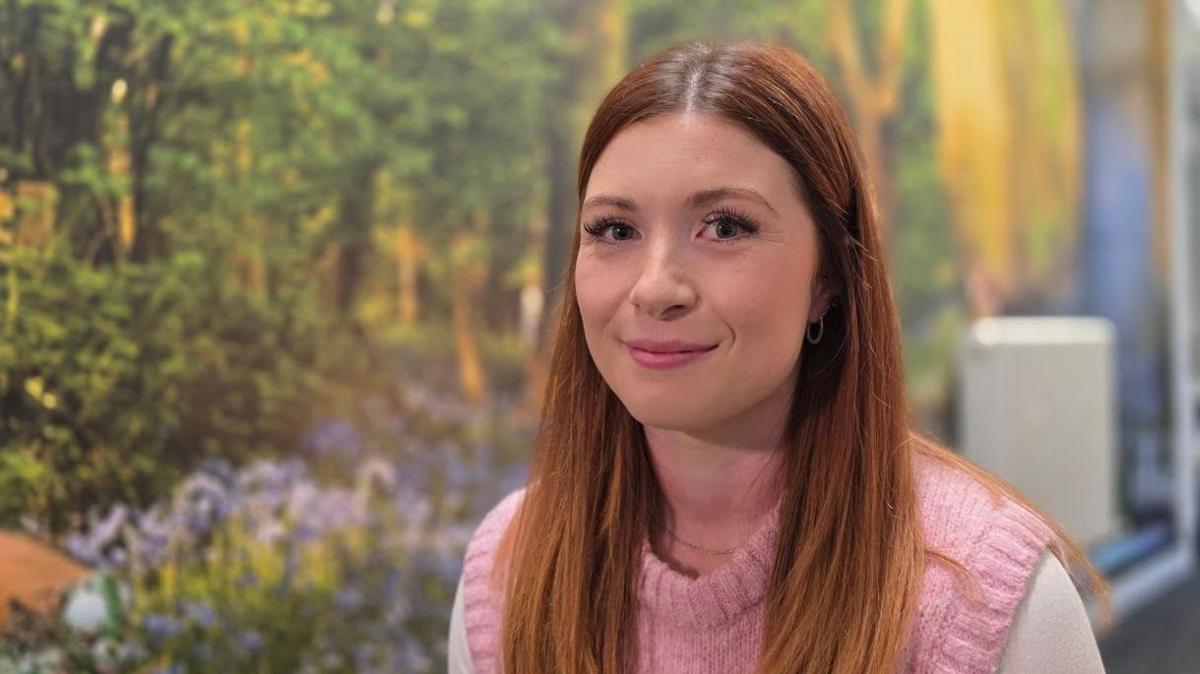 Nina Finnergan is sitting in front of a wall decorated to look like a forest. She has pink tank-top on top of a white shirt.