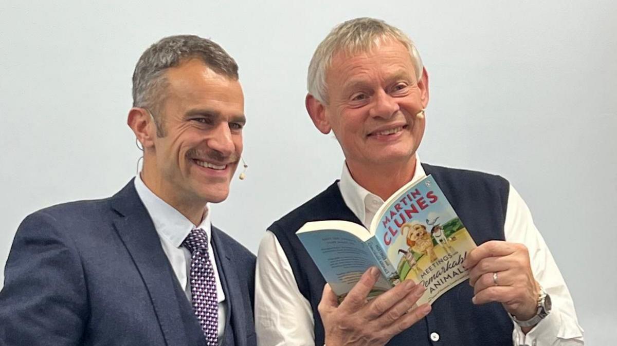 Steve Harris and Martin Clunes smile for the cameras at the Dorchester Literary Festival. Steve Harris, on the left, is wearing a navy suit, white shirt and checkered tie. He has dark hair and a moustache. Martin Clunes is holding up a paperback copy of his book - Meeting Remarkable Animals.