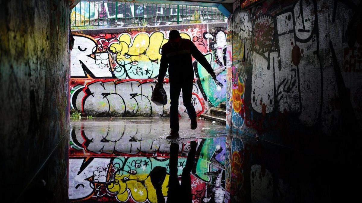 A person shelters from the rain in the flooded Lawrence Hill underpass in Bristol. They are silhouetted against the wall behind, and are carrying a bag in one hand. Behind them is a wall covered in graffiti, and the person and the graffiti is reflected in the water in the underpass