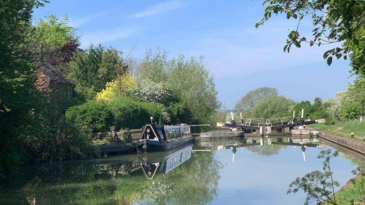 The canal at Southam. A narrowboat is reflected in the calm water on the left as it lies moored up. On the right is a partly closed lock gate. Trees and grass line the canal on the left and right