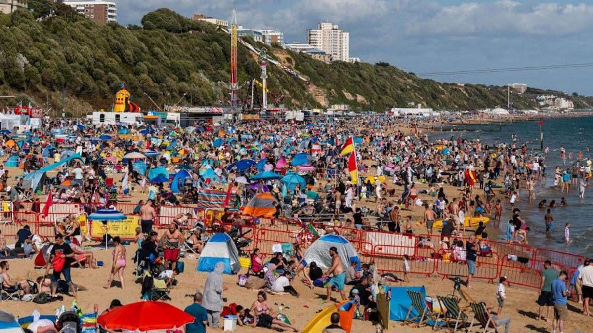 A packed Bournemouth beach with funfair rides and sloping cliffs covered with vegetation in the background