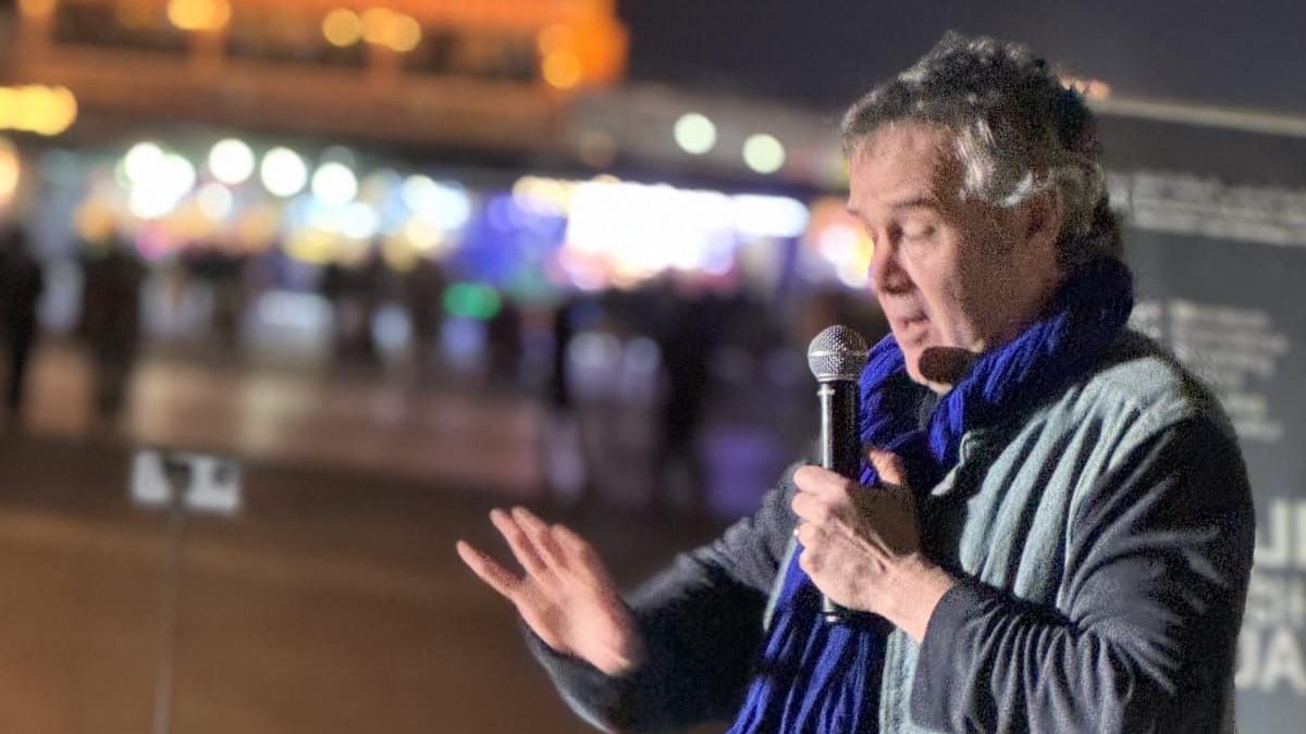 Maxwell speaking at the festival. He wears a blue and grey jacket and an electric blue scarf, and is speaking into a microphone. 