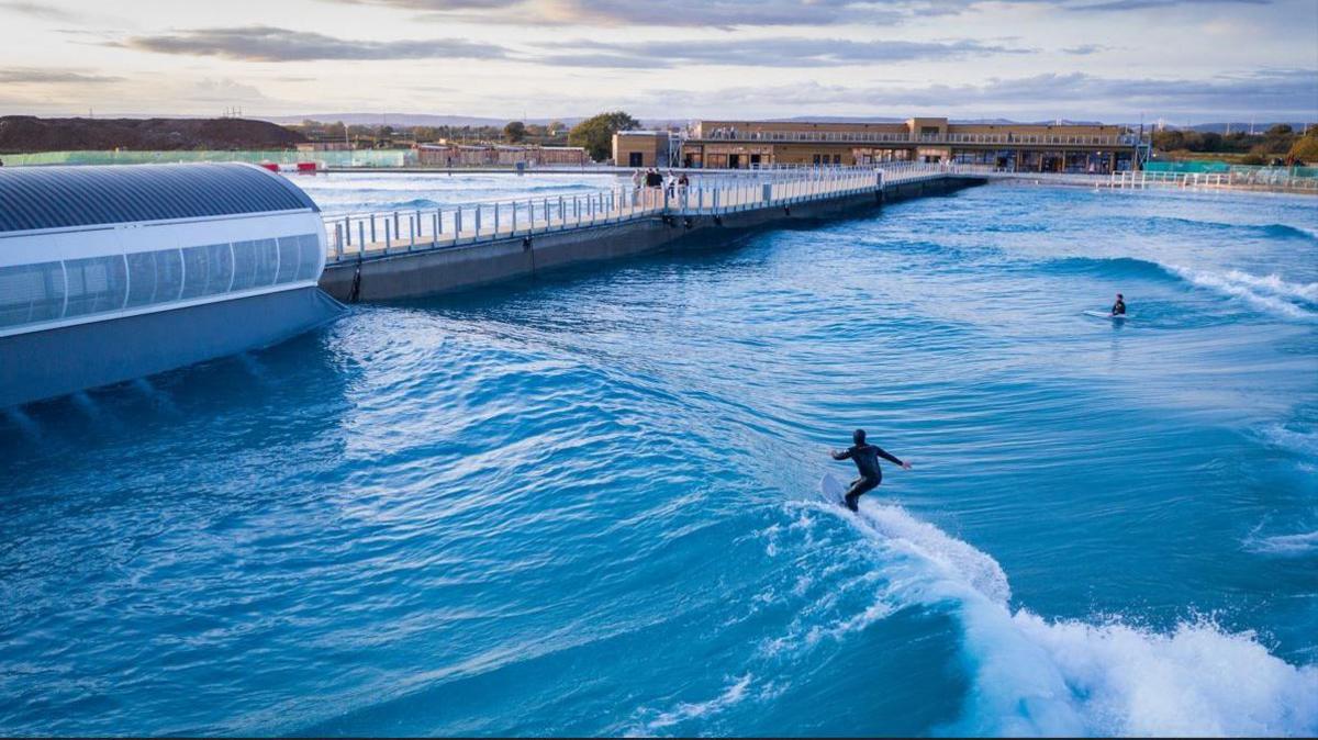 An aerial picture taken from above the water at The Wave. It appears to be dusk, and the water is a deep vibrant blue. In the middle of the water is a wooden pier for spectators to watch. There are two surfers in the water on the right, and one is standing up riding a wave. 