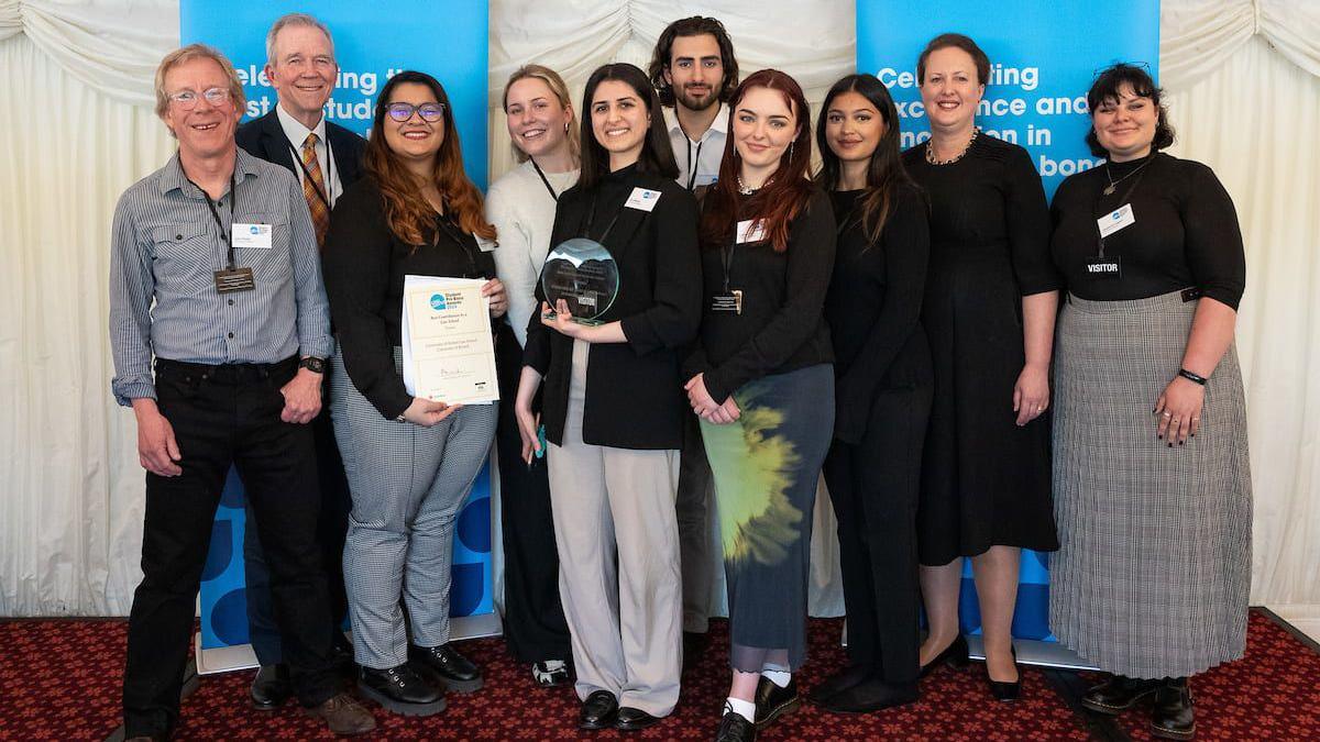 A group of 10 people - seven women and three women - stand smiling as the face the camera in a room with a dark red carpet. Behind them is a set of white drapes and two light blue banners.