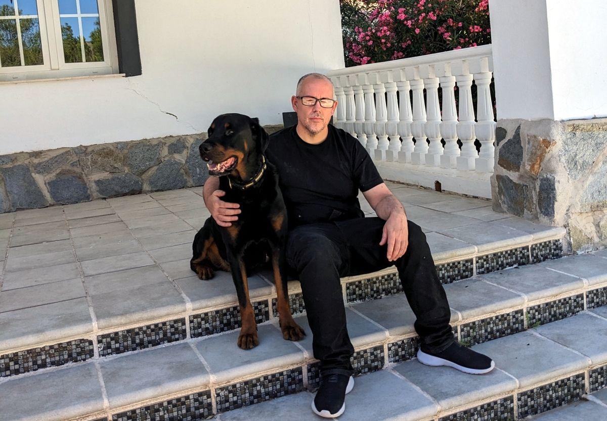 Ryan O'Meara sitting on a step outside a house with his dog. He wears a dark t-shirt and jeans and black trainers with white soles. He is wearing glasses. His dog is large, brown and black and with big teeth. 