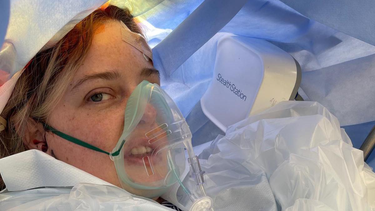 Sophie lies on a surgical table covered in a blue drape, wearing a clear oxygen mask. Medical equipment labeled "StealthStation" is visible in the background, indicating a clinical or surgical environment