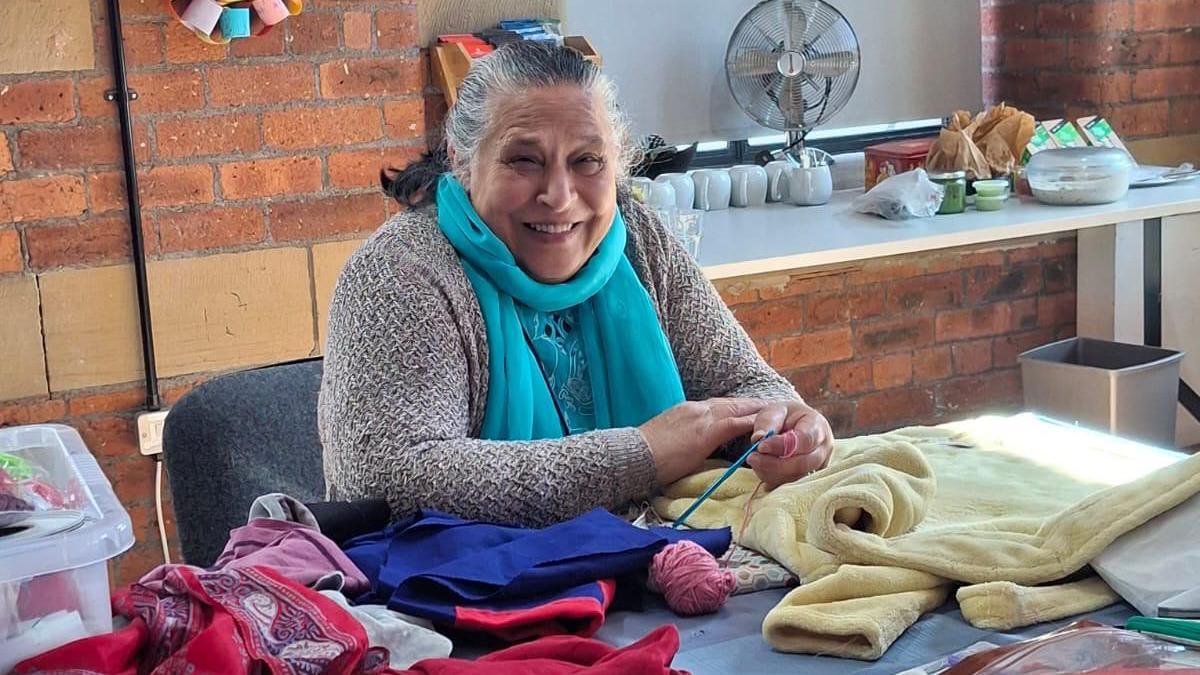 A woman, possibly in her 60s or early 70s, sits with a broad smile behind table which is full of multicoloured bits of material. She is holding some knitting needles and has a ball of wool in front of her.