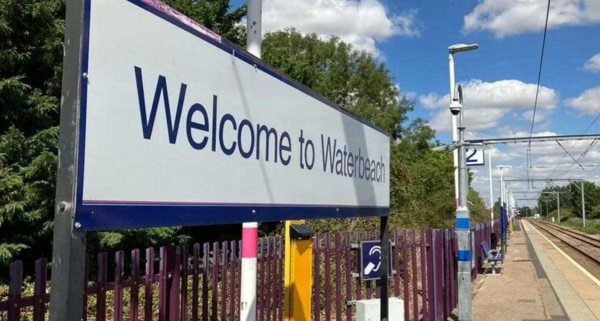 A sign saying 'Welcome to Waterbeach' above purple railings and a train platform and track to the right. 