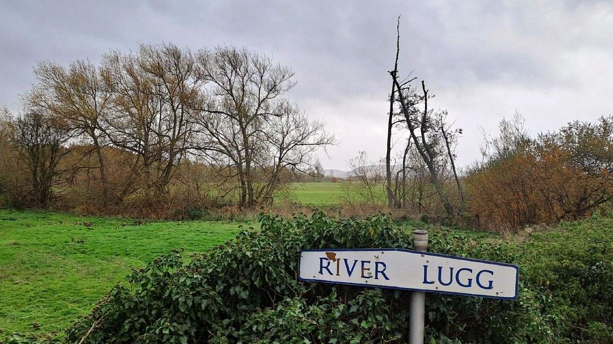 In the foreground is a sign which reads "River Lugg" while in the background is a meadow with trees and bushes.