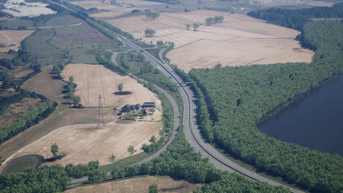 An aerial view of the road running through a series of fields and a wooded area. There is a body of water to the bottom right corner of the image.