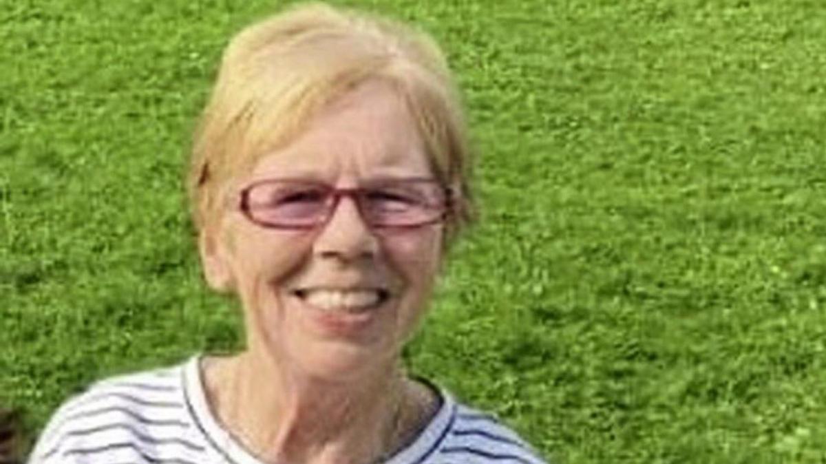 Una Noone is smiling into the camera. She has glasses on and a black and white stripped top. She had short blond hair. She's standing in a field. 