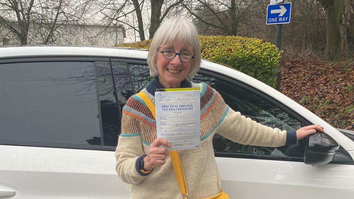 An older woman with blue glasses holds up a driving certificate and stands in front of a white car.