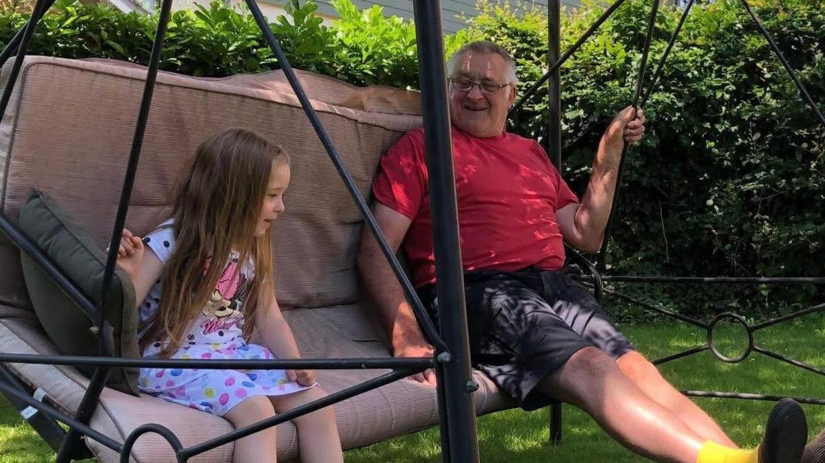 Lucy, on the left, and her Grandad Easton, on the right, are sat on an outside swing which has is black with a beige cushion. Behind them is a hedge which goes around the image.