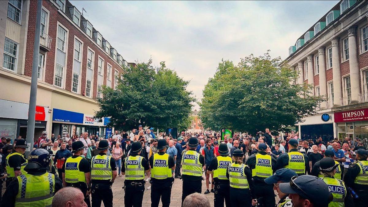 Police officers stand in a line in King Edward Street in Hull as a group of protestors head towards them. The officers are wearing high-visibility vests and helmets.