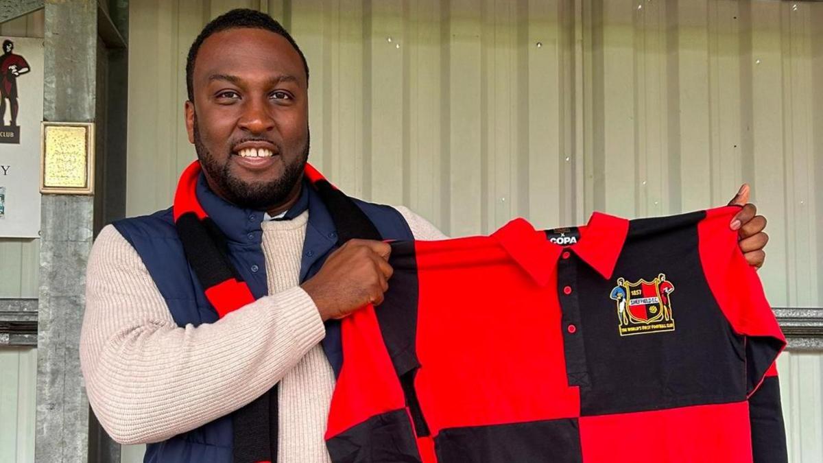 Sheffield FC manager Vill Powell, who is holding a red and black-coloured Sheffield FC shirt. Mr Powell is a black man with short black hair and short-length facial hair.