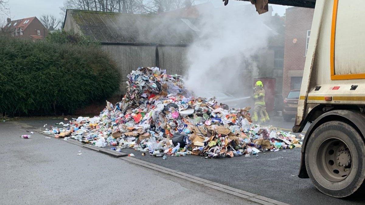 The pile of rubbish dumped in a pub car park. The rubbish is smouldering. A firefighter is training a hose on it. The back part of the bin lorry is also in the picture