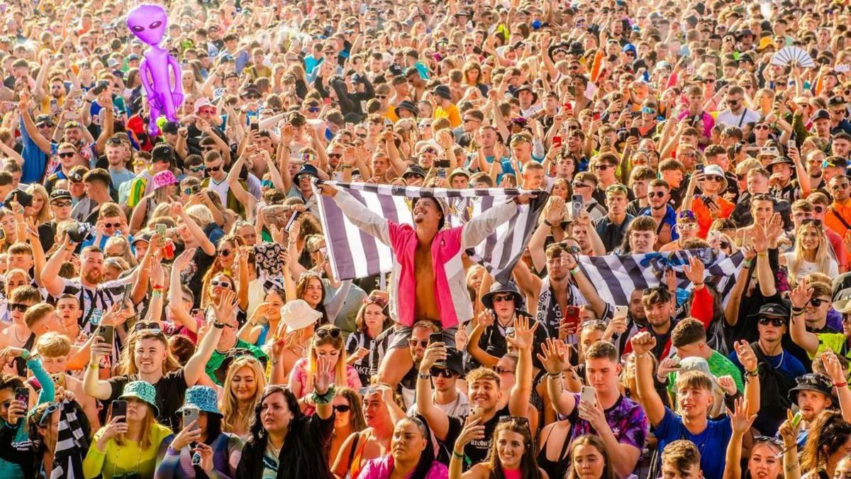 Crowds at last year's festival hold their hands in the air. One man holds up a black and white flag while wearing a pink shirt and his head tips back smiling. There is a purple alien balloon behind him. Some in the crowd hold up mobile phones and are wearing bucket hats.