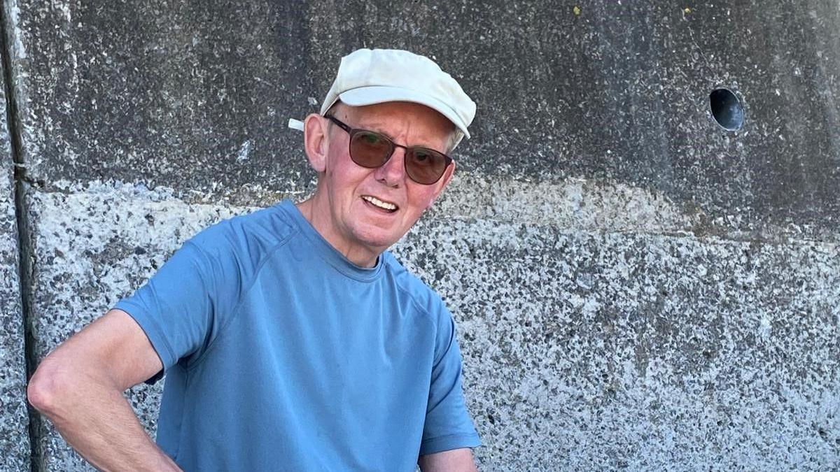 A man wearing a pale cap, a blue tshirt and sunglasses in front of a weathered concrete wall
