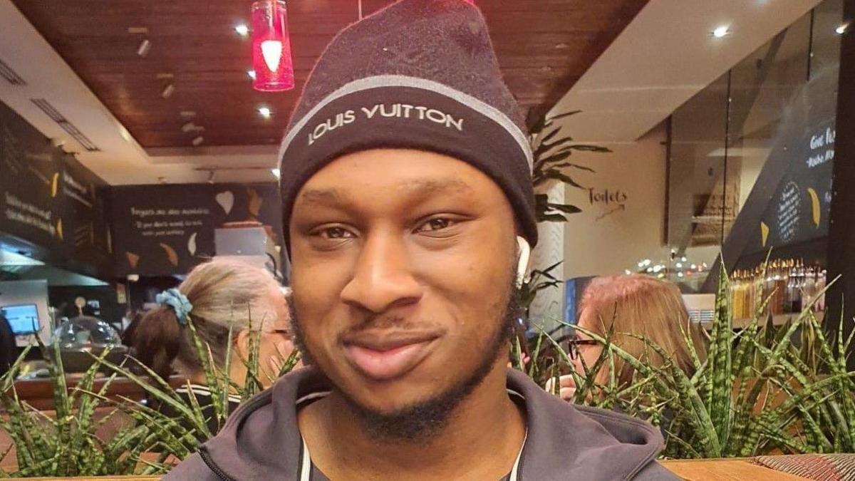 Khaleed Oladipo wearing a black hat and zip-up jacket, smiling.