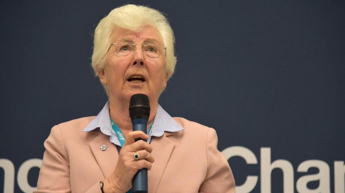 Doncaster Mayor Ros Jones: she is wearing a dusky pink blazer and pale blue shirt and talking into a microphone in her right hand.  She has short white hair and wears thin-rimmed glasses.  She has a pin on her lapel and is wearing a gemstone ring on her right hand.