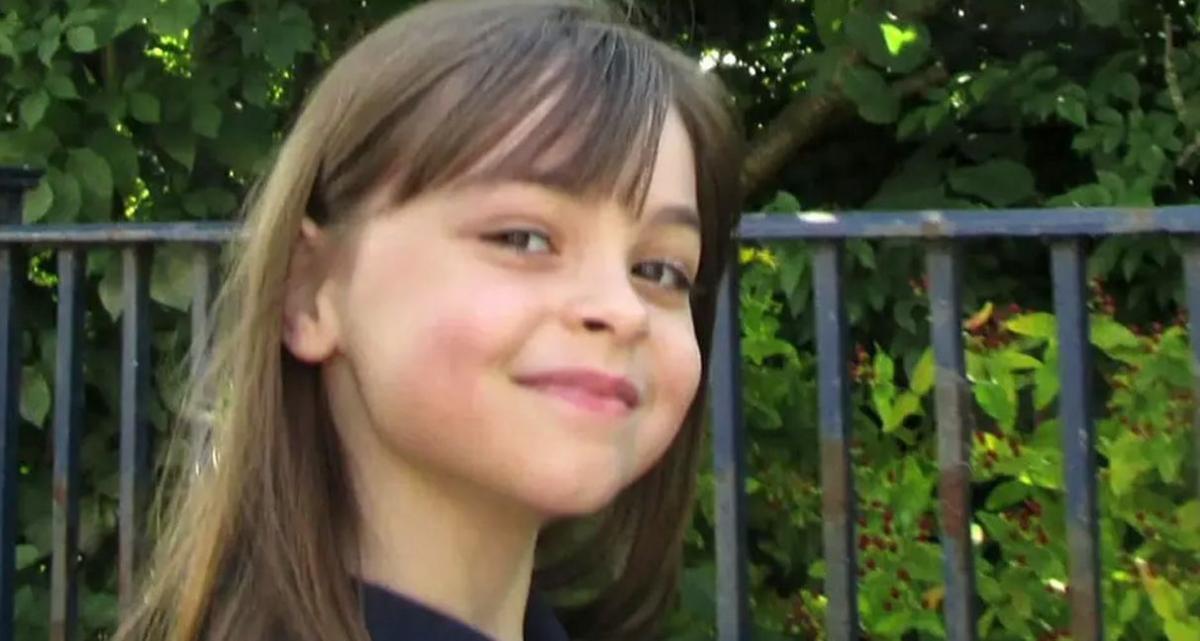 Saffie Roussos standing in front of greenery with a cheeky smile on her face. She has long brown hair with a short fringe