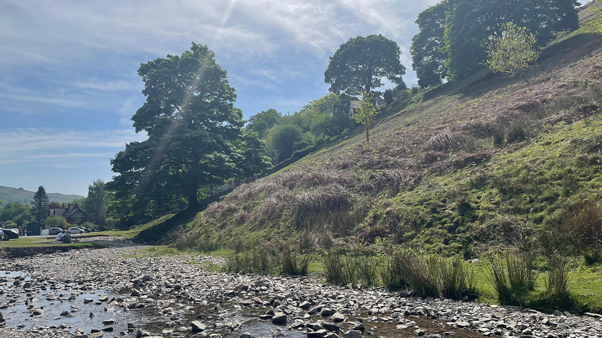 A shallow stream near Church Stretton with a steep hill rising up to the right, covered in bracken and grass with a scattering of trees. A car park is in the distance on the left