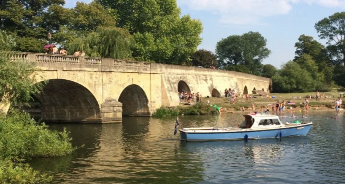 Wallingford Beach in Oxfordshire with a blue and white boat on the river and a bridge in the background