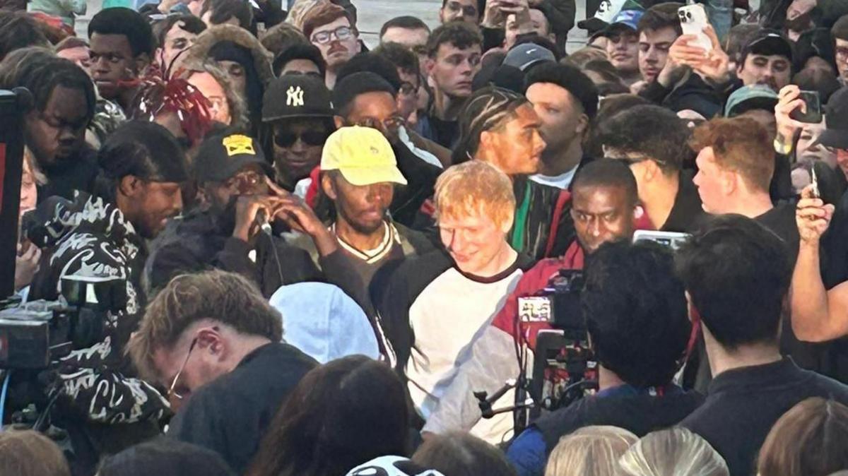 A ginger-haired man wearing a white t-shirt with black markings is surrounded by people.